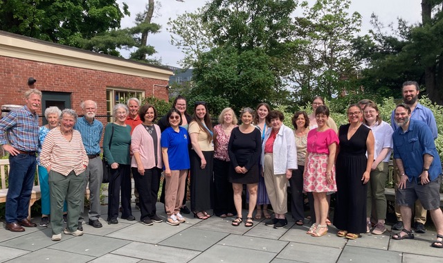 A group of 20 people standing on a stone-paved patio in front of a red brick building and lush greenery.