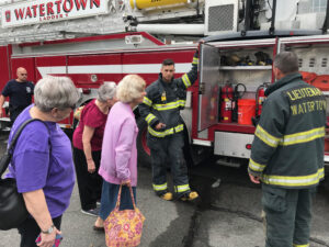 A group of elderly women is inspecting the open compartment of a fire truck, guided by two firefighters. The fire truck has "Watertown Ladder 1" written on its side, and one firefighter is holding the compartment door open while another stands nearby, facing away from the camera.