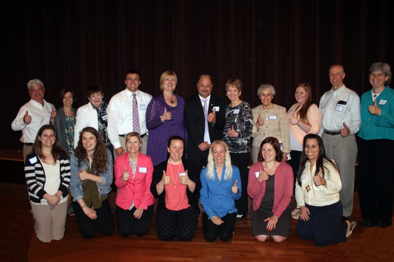 Group of sixteen people posing with thumbs up in front of a dark curtain.