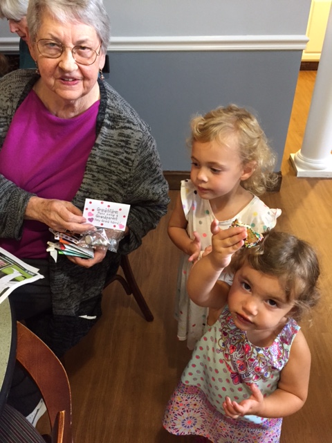 An elderly woman with glasses is sitting and holding a card, smiling. Two young girls with curly hair stand next to her