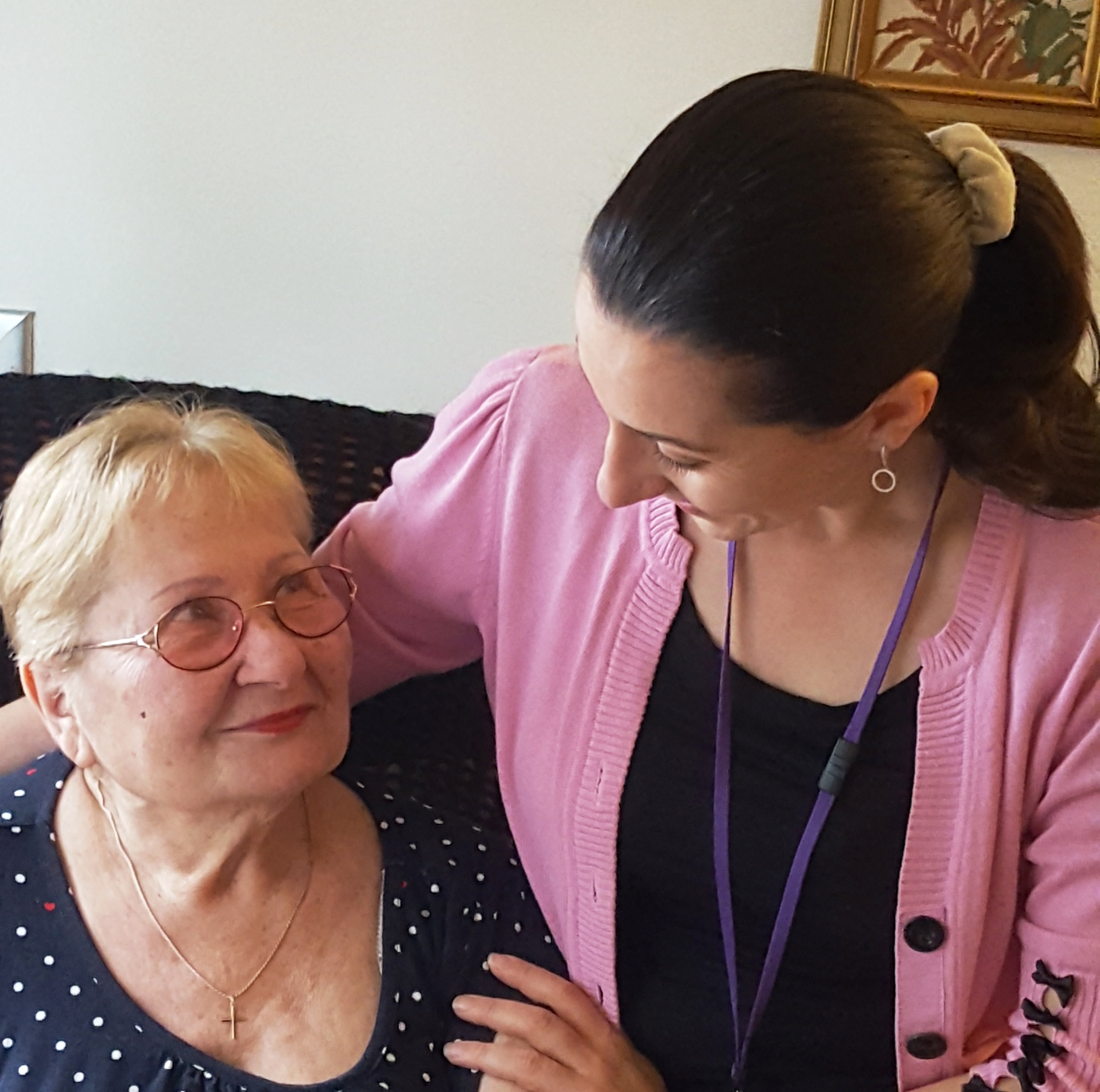 Two women sitting close together, an older woman with glasses and short grey hair smiling at the camera, and a younger woman with dark hair in a ponytail wearing a pink cardigan looking at the older woman affectionately.