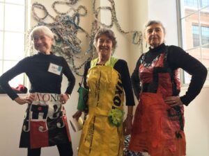 Three senior women standing together, smiling and posing confidently, wearing colorful aprons made from recycled materials. Behind them is a wall-mounted artwork made of woven or twisted materials.