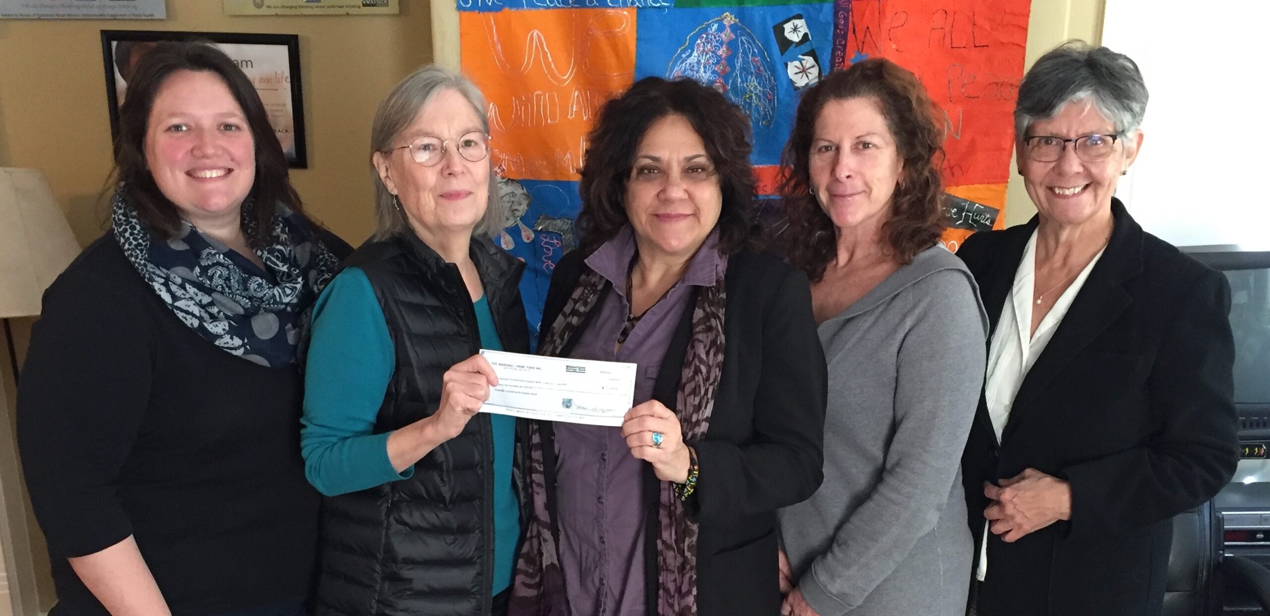 Five women standing together, smiling at the camera. Two women in the center are holding a check. Colorful artwork is visible on the wall behind them.