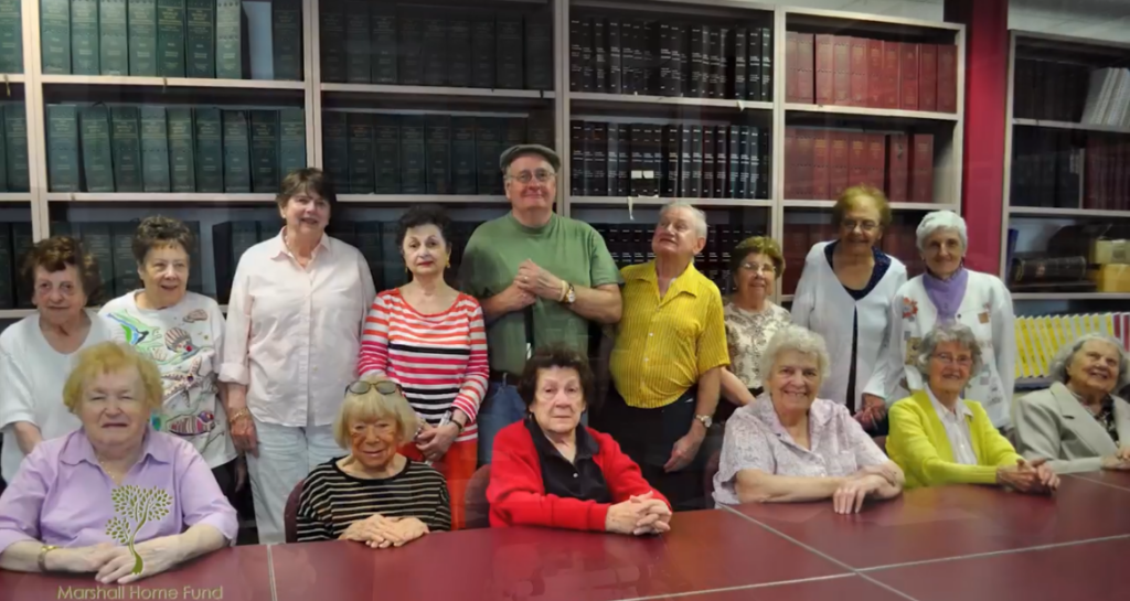 A cheerful group of seniors poses in front of a table in the Watertown Library for the WCA-TV Public Access PSA