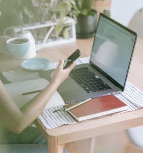 A cozy workspace with a laptop computer, 2 paper notebooks, & coffee cup on a table. A person holds a smartphone.